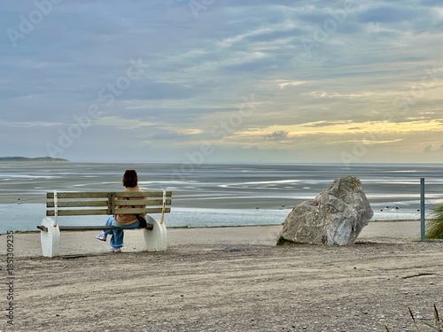 Woman sitting on bench overlooking Baie d'Authie at low tide in Berck, France, tranquil seascape with sandbanks, clouds and soft sunset light, solitude and travel