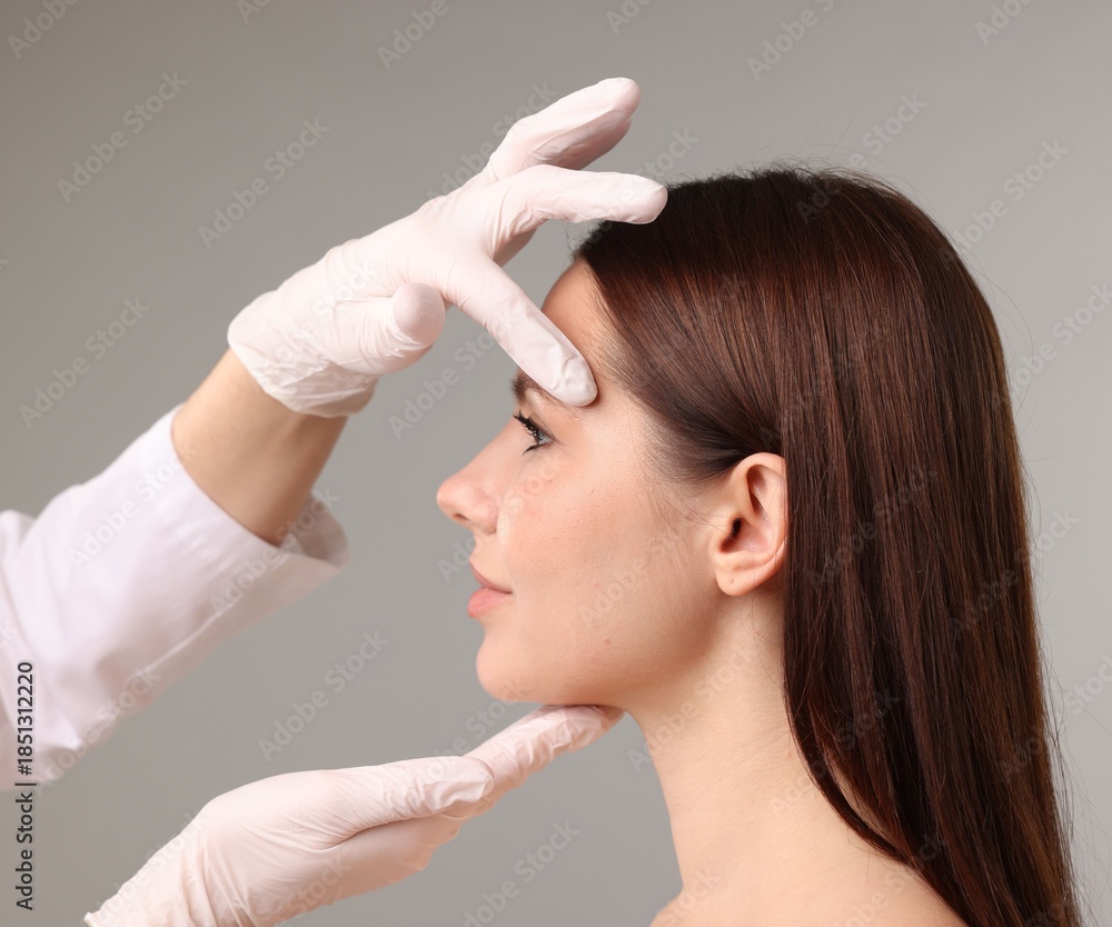 Fototapeta premium Doctor checking patient's face before plastic surgery operation on grey background, closeup