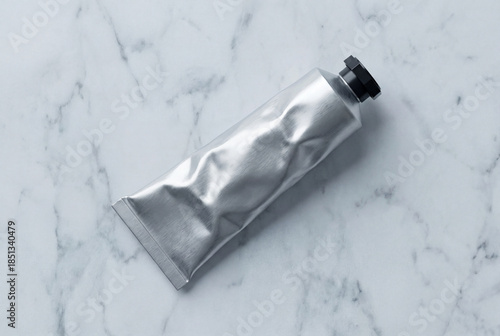Top down flat lay of blank matte aluminum squeeze tube mockup with black octagonal cap on white marble surface with realistic crinkle texture
