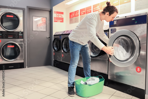 Obraz na plátně man loading clothes into washing machine at self-service laundromat with green basket