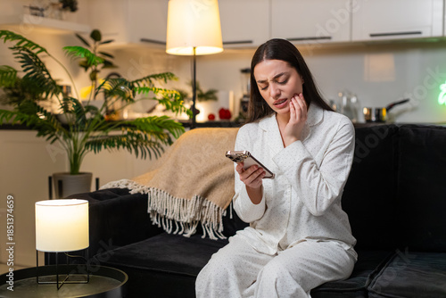 Foto Young woman at night home holds cheek tooth pain uses smartphone to find dentist book visit