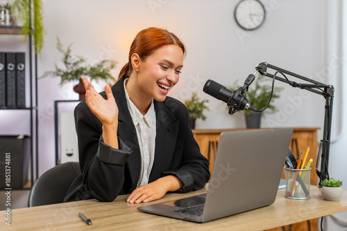 Young businesswoman at home office talks into studio microphone recording podcast blog video laughing. Freelancer girl by table near laptop shares ideas engaging audience and enjoying fun joyful work.