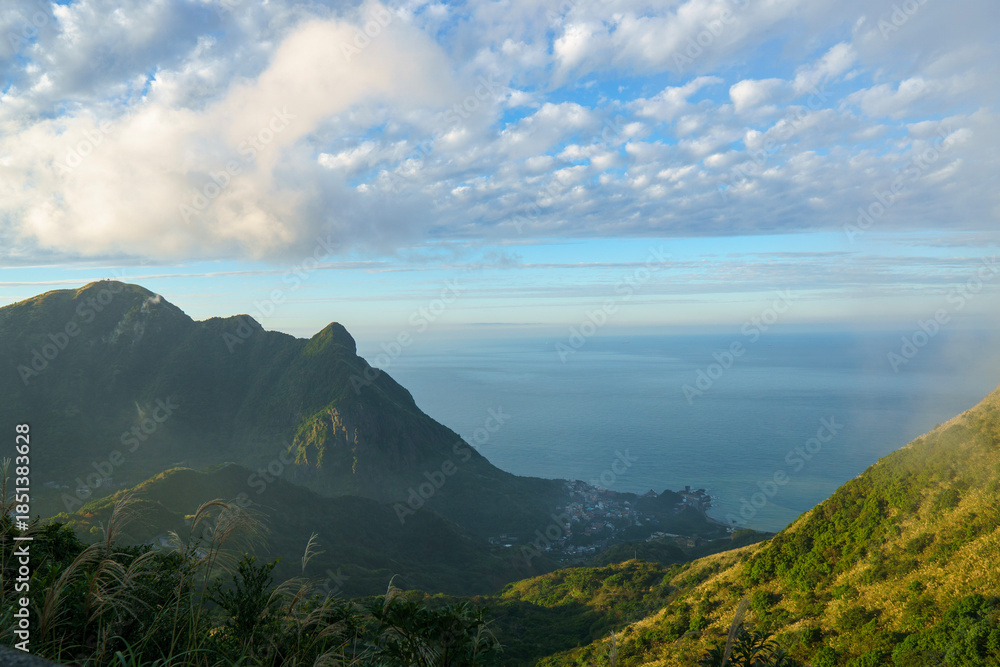 Fototapeta premium A wide view of a green mountain valley opening toward the sea under a blue sky, capturing peaceful nature, landscape depth, and outdoor travel scenery.