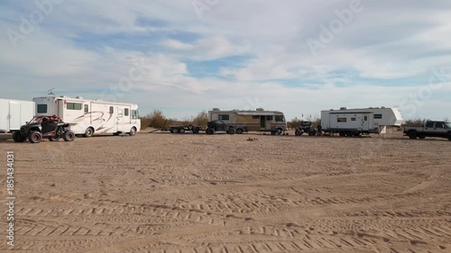 Aerial orbit of a desert camp with motorhomes, trailers, and UTVs parked in a circle.