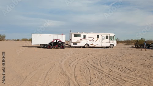 Aerial orbit of a desert camp with motorhomes, trailers, and UTVs parked in a circle.