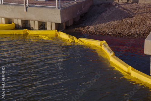 Industrial Waterfront With Yellow Oil Boom water goat Barriers Along Calm Water Near Concrete Pier Structure. Floats along the water beside a concrete pier, illustrating spill response equipment at an