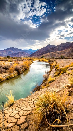 River winds through a dry, cracked landscape under a dramatic sky of clouds and mountains
