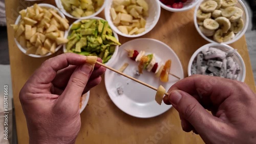 Close-up of men's hands stringing pieces of fresh fruit on a wooden skewer to make fruit kebabs