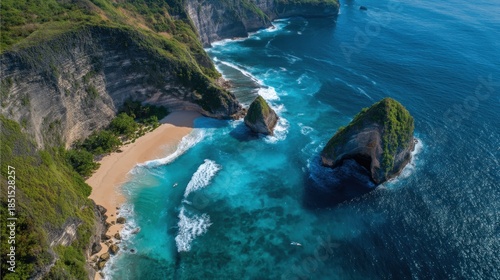A beach is seen surrounded by high cliffs and large rock formations. Waves crash on the shore. The sun shines brightly on the blue water. This scene shows a tropical coastal area.