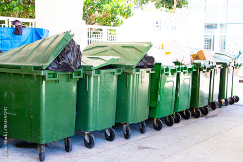 Old rubbish bin large size garbage square shape green with flip cover arranged in same pattern. Old dirty trash black cans plastic. Waste collection equipment in park. Protect environment.