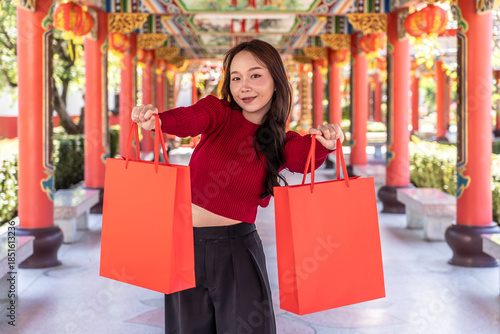 Portrait of joyful young asian woman celebrates Chinese New Year at chinese temple, showcasing her bright red shopping bags against a stunning shrine backdrop. A moment of happiness