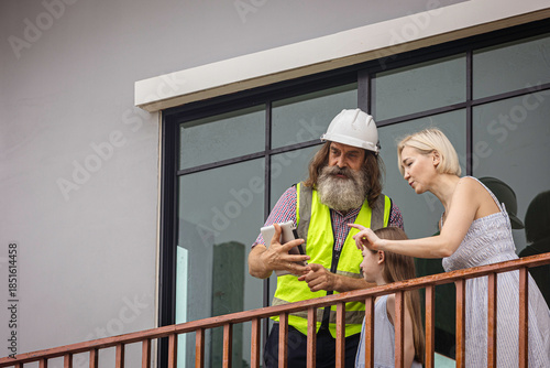 Experienced bearded foreman shows new home plans on a tablet to a smiling mother and daughter over terrace, highlighting smart construction technology and a warm, hopeful future in their house