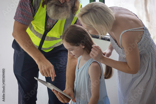 Experienced bearded foreman shows new home plans on a tablet to a smiling mother and daughter, highlighting family trust, smart construction technology and a warm, hopeful future in their house