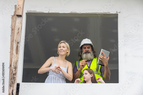 Experienced bearded foreman shows new home plans on a tablet to a smiling mother and daughter over terrace, highlighting smart construction technology and a warm, hopeful future in their house