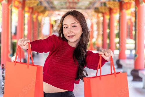 Portrait of joyful young asian woman celebrates Chinese New Year at chinese temple, showcasing her bright red shopping bags against a stunning shrine backdrop. A moment of happiness