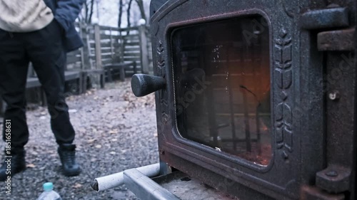 Bright orange flames burn vigorously inside a rustic cast iron stove with a glass window. A person wearing winter boots and dark trousers stands on the gravel nearby.