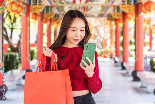 An Asian woman joyfully explores online shopping at a colorful Chinese temple during Lunar New Year. Holding red bags, she embraces festive vibes while connecting digitally.