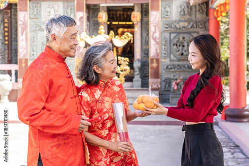 During the Lunar New Year at a beautiful Chinese temple, a cheerful teenage daughter presents her parents with oranges, symbolizing love and respect. Together, they cherish this festive tradition.