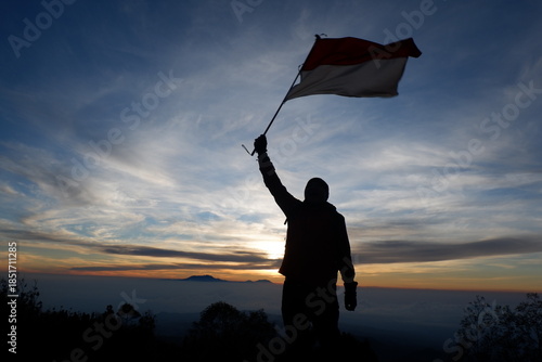 Silhouette of a man standing while carrying the Indonesian red and white flag with the sky and mountainous horizon.
