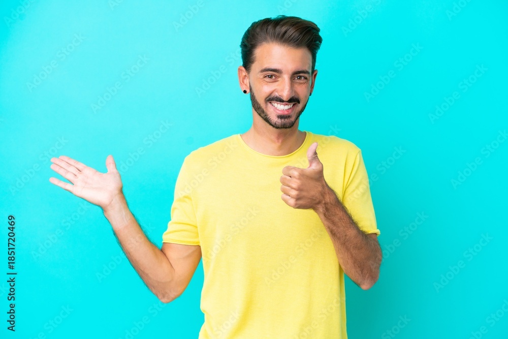 Fototapeta premium Young caucasian man isolated on blue background holding copyspace imaginary on the palm to insert an ad and with thumbs up