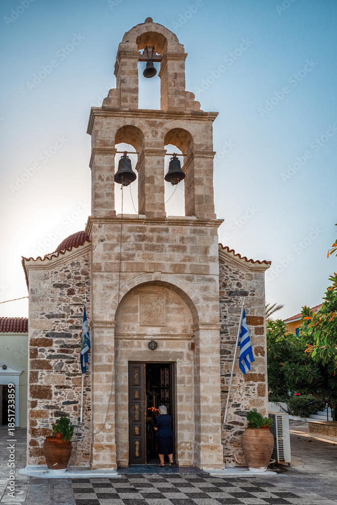Fototapeta premium Stone church with bell tower in Panormos village on Crete featuring traditional architecture and Greek flags