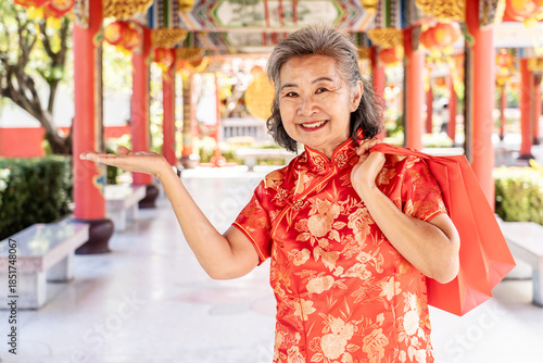 A happy senior Asian woman dressed in a bright red cheongsam holds festive shopping bags while beaming at a beautiful Chinese temple during Lunar New Year celebrations, festive shopping, tradition