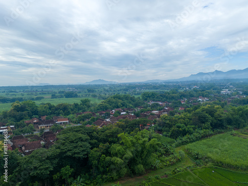 Peaceful aerial view of a traditional rural village and rice fields surrounded by lush greenery and distant lawu mountain under a cloudy sky. Residential area in the tropics.