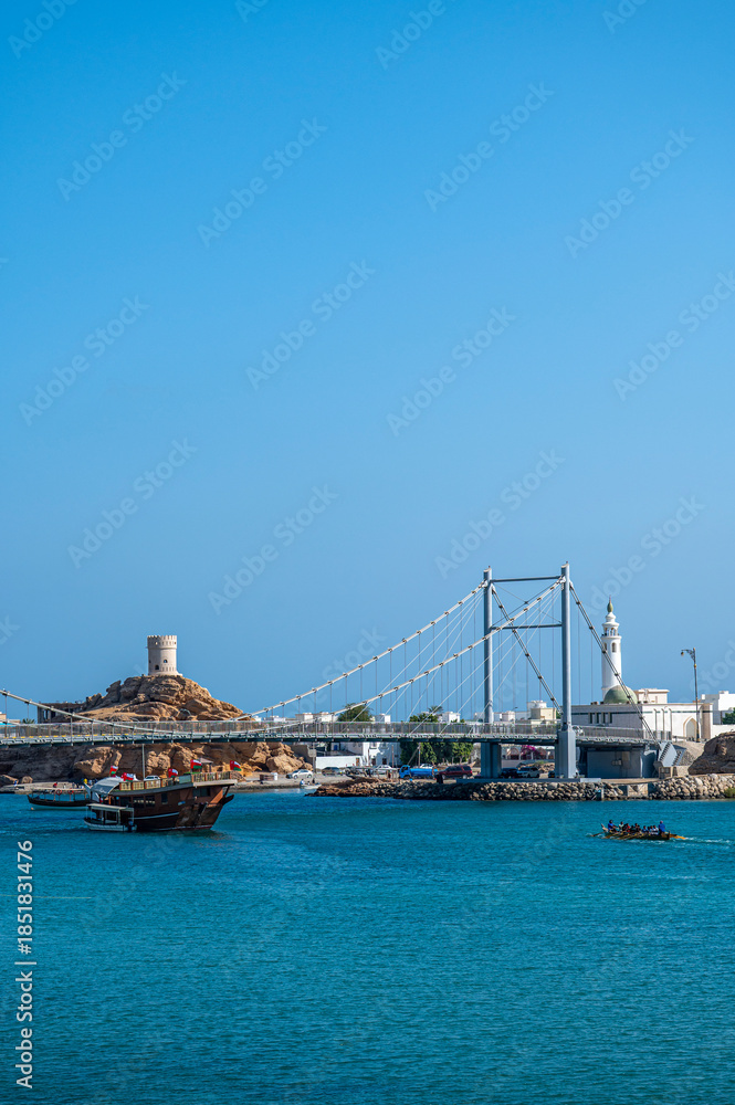 Fototapeta premium Sur, Oman - November 22, 2025: Suspension Bridge, Historical Watchtower, and Mosque Minaret, Sur, Oman