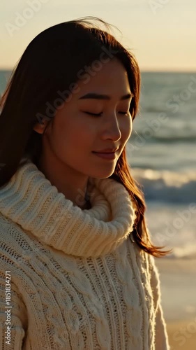 profile of beautiful asian woman relaxing on beach during sunset. girl wearing warm white knitted sweater. ocean background with golden light. vacation, mental health concept.