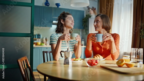 Photos Two besties holding cups with hot tea sitting together at apartment kitchen