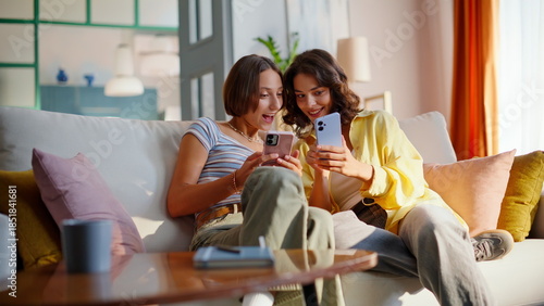Laughing besties holding cellphones sitting cozy home couch. Smiling girlfriends