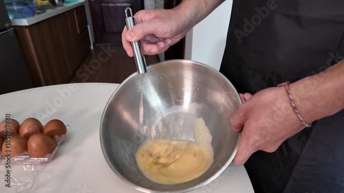 A man is beating eggs with a whisk in a metal bowl on the kitchen table, a man is wearing a black apron