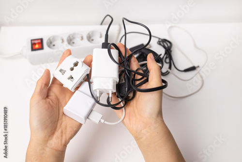 Hands holding various electrical appliances plugs near white extension cord. Electrical overload. Overloaded power plugs, close up