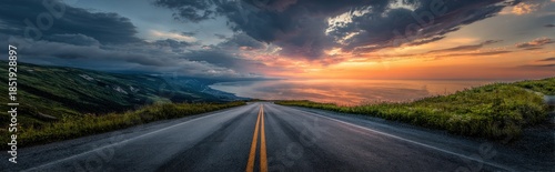 A serene asphalt road stretches through a lush valley at sunset with dramatic clouds.