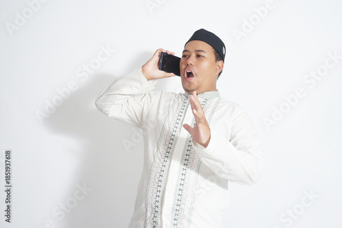 An Indonesian Muslim man wearing a black peci and a white koko shirt is shocked while holding a cellphone with white background isolated.