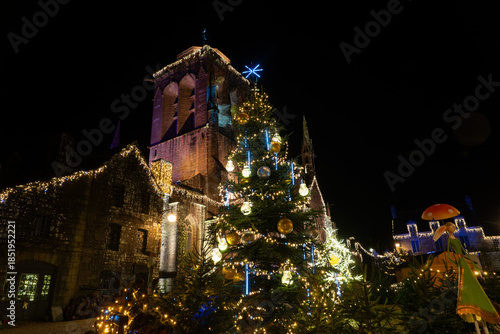 Église du village de Locronan en Bretagne illuminée pour les fêtes de Noël, sapin décoré et décorations lumineuses créant une ambiance nocturne chaleureuse et festive.