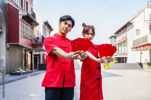 Asian man and woman proudly display bright red envelopes while wearing stylish cheongsam attire. They celebrate Chinese New Year together in a cheerful atmosphere, Female and Male show Ang Pao