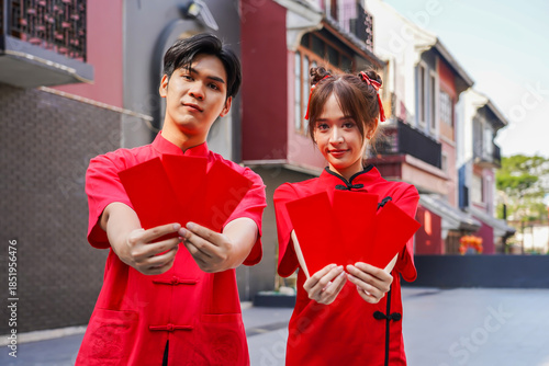 Asian man and woman proudly display bright red envelopes while wearing stylish cheongsam attire. They celebrate Chinese New Year together in a cheerful atmosphere, Female and Male show Ang Pao