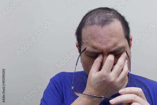 An Asian man in a blue shirt holds his glasses while rubbing the bridge of his nose and eyes, showing signs of digital eye strain, exhaustion, vision fatigue, migraine, or stress