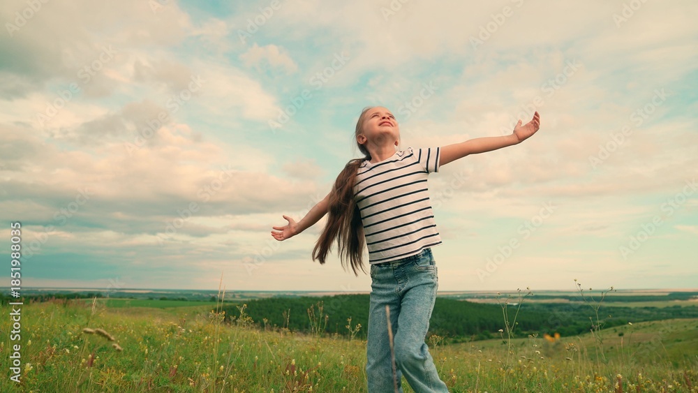 Fototapeta premium Kid girl plays on lawn under sky, childhood dream travel. Child girl enjoys green nature in summer, raising her arms. Kid in park. Slow motion. Little girl plays in meadow, dream of fly. Happy family