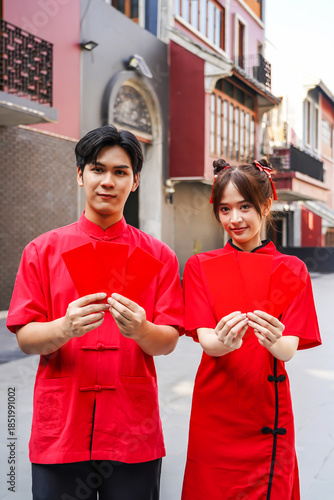 Asian man and woman proudly display bright red envelopes while wearing stylish cheongsam attire. They celebrate Chinese New Year together in a cheerful atmosphere, Female and Male show Ang Pao