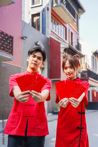 Asian man and woman proudly display bright red envelopes while wearing stylish cheongsam attire. They celebrate Chinese New Year together in a cheerful atmosphere, Female and Male show Ang Pao