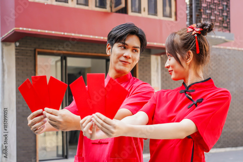 Asian man and woman proudly display bright red envelopes while wearing stylish cheongsam attire. They celebrate Chinese New Year together in a cheerful atmosphere, Female and Male show Ang Pao