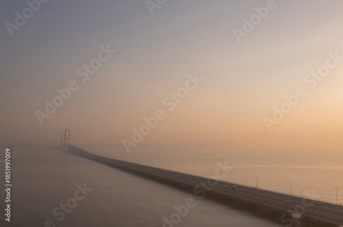 Aerial view of the long Suramadu Bridge stretching across the ocean during a hazy sunrise, with the bridge structure fading into the golden misty horizon