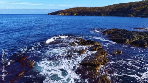 The sun rises over a serene coastline, illuminating dark rocks as waves crash rhythmically, sending white foam into the air amidst a backdrop of calm sea and distant hills.