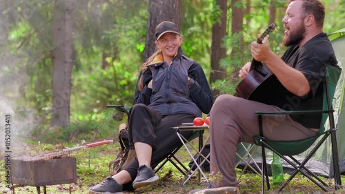 Happy young couple enjoying a picnic in the forest, where a bearded man plays the guitar and sings for his smiling girlfriend while meat cooks on the grill next to their tent