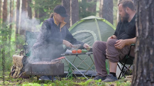 Young couple enjoying a romantic picnic at their campsite in the woods while the bearded man plays the guitar for his smiling girlfriend near the tent and smoking campfire on a sunny day
