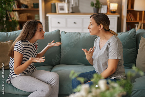 A teenage girl expresses frustration while sitting on a couch, facing her mother who responds calmly. The atmosphere reflects a tense yet loving family moment.