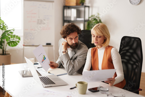 Two team members sit at a table in a bright office. They look at charts and graphs on paper while discussing strategies and promoting success together.