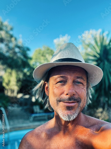 Happy blond man with straw hat and a stubble beard by a pool in a sunny summer garden.